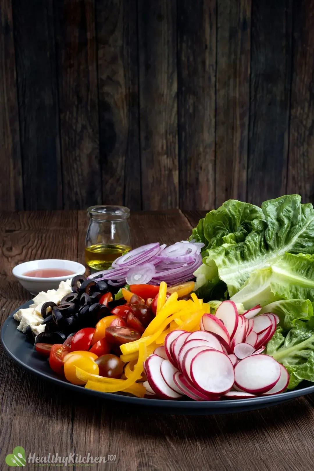 A large plate of vibrant and colorful vegetables for a salad: radishes, pepper, lettuce, tomatoes, olives, red onions, and feta