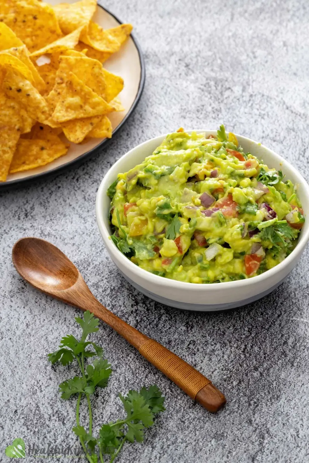 A close-up of guacamole with diced tomatoes and onions, garnished with cilantro on a white plate, next to a wooden spoon and a plate of chips