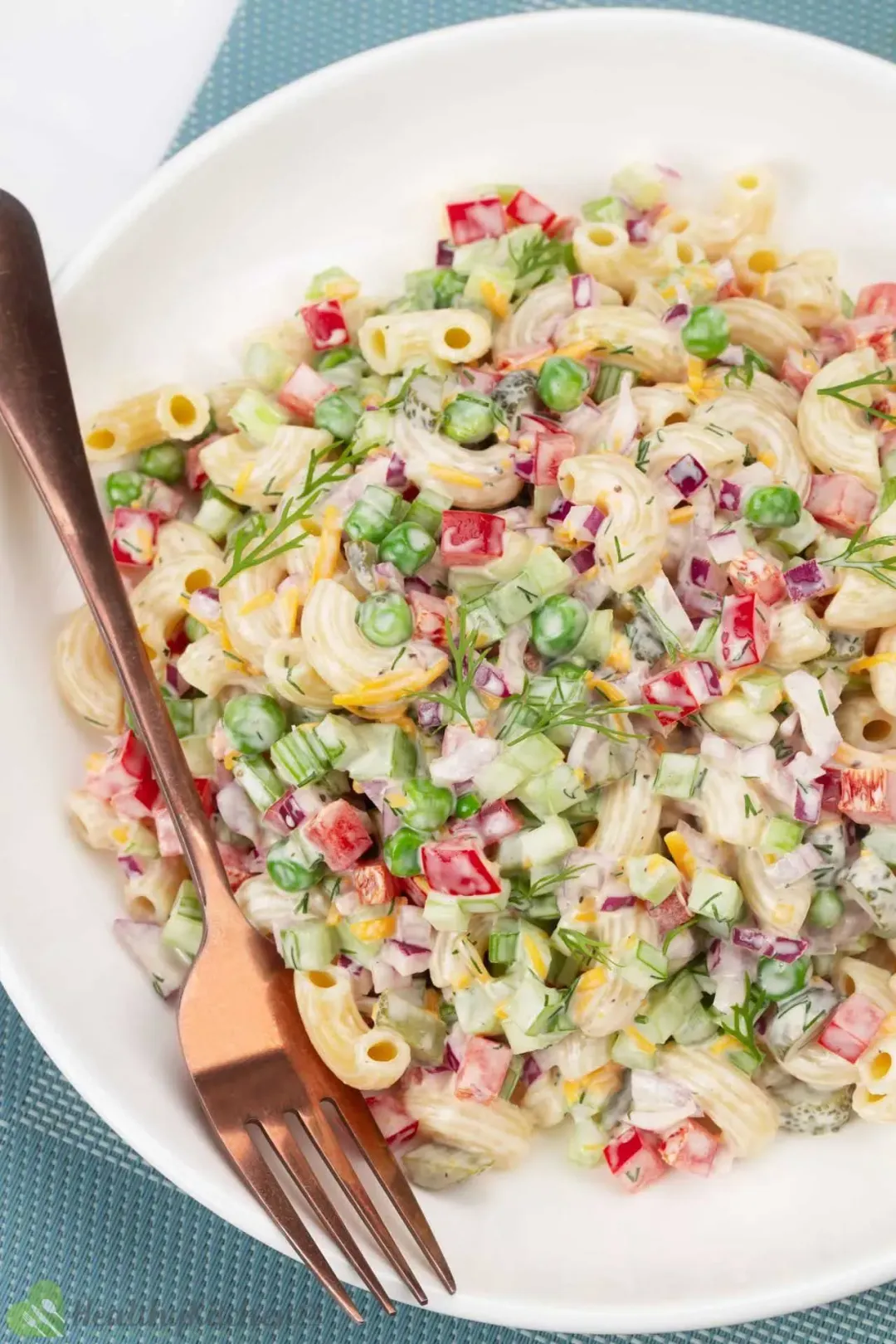 A white plate of macaroni salad with bits of peas, cheese, dill, pepper, and red onion, with a golden fork
