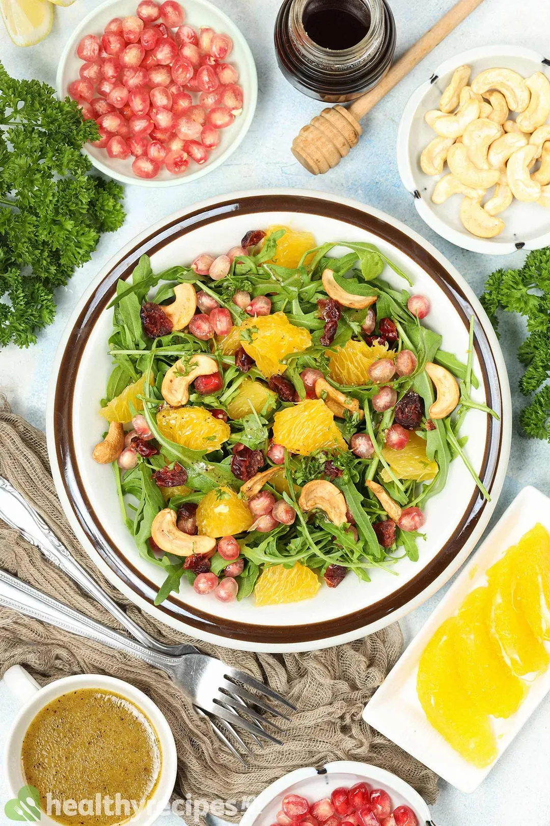 A high-angle shot of a plate of arugula salad placed near a spoon, a fork, plates of pomegranate seeds, cashews, and orange segments.