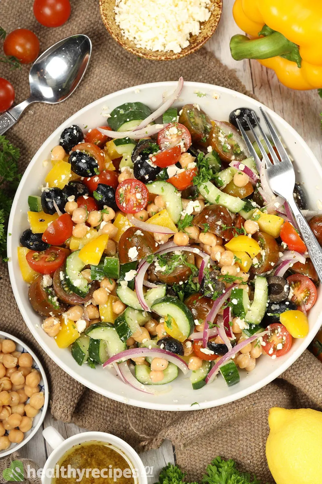 A round white plate containing Greek chickpea salad with a fork on the side, laid nearby is a spoon, cherry tomatoes, a disk of chickpeas, a yellow bell pepper, and a small dressing bowl.