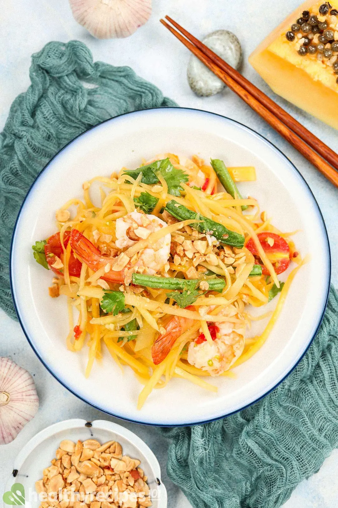 A plate of papaya salad laid on a blue mesh cloth near chopsticks and roasted peanuts