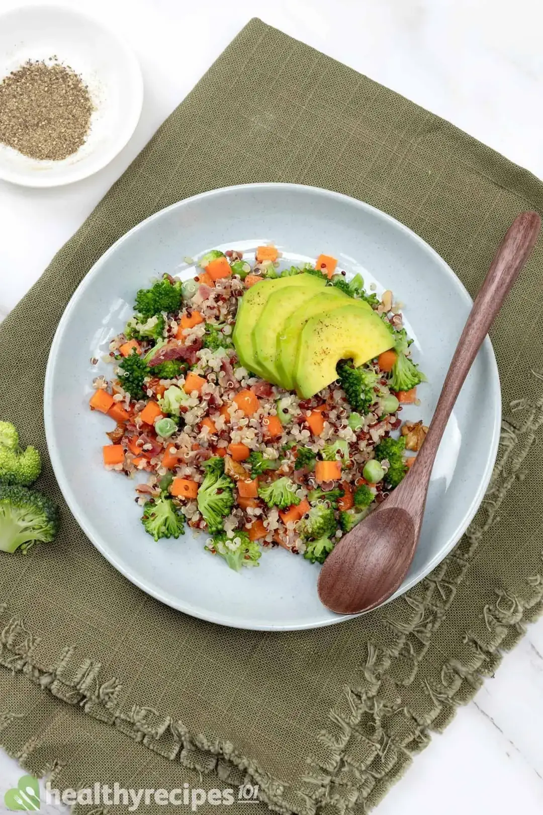 A plate of quinoa salad with avocado slices in a white plate on a green kitchen cloth with a wooden spoon