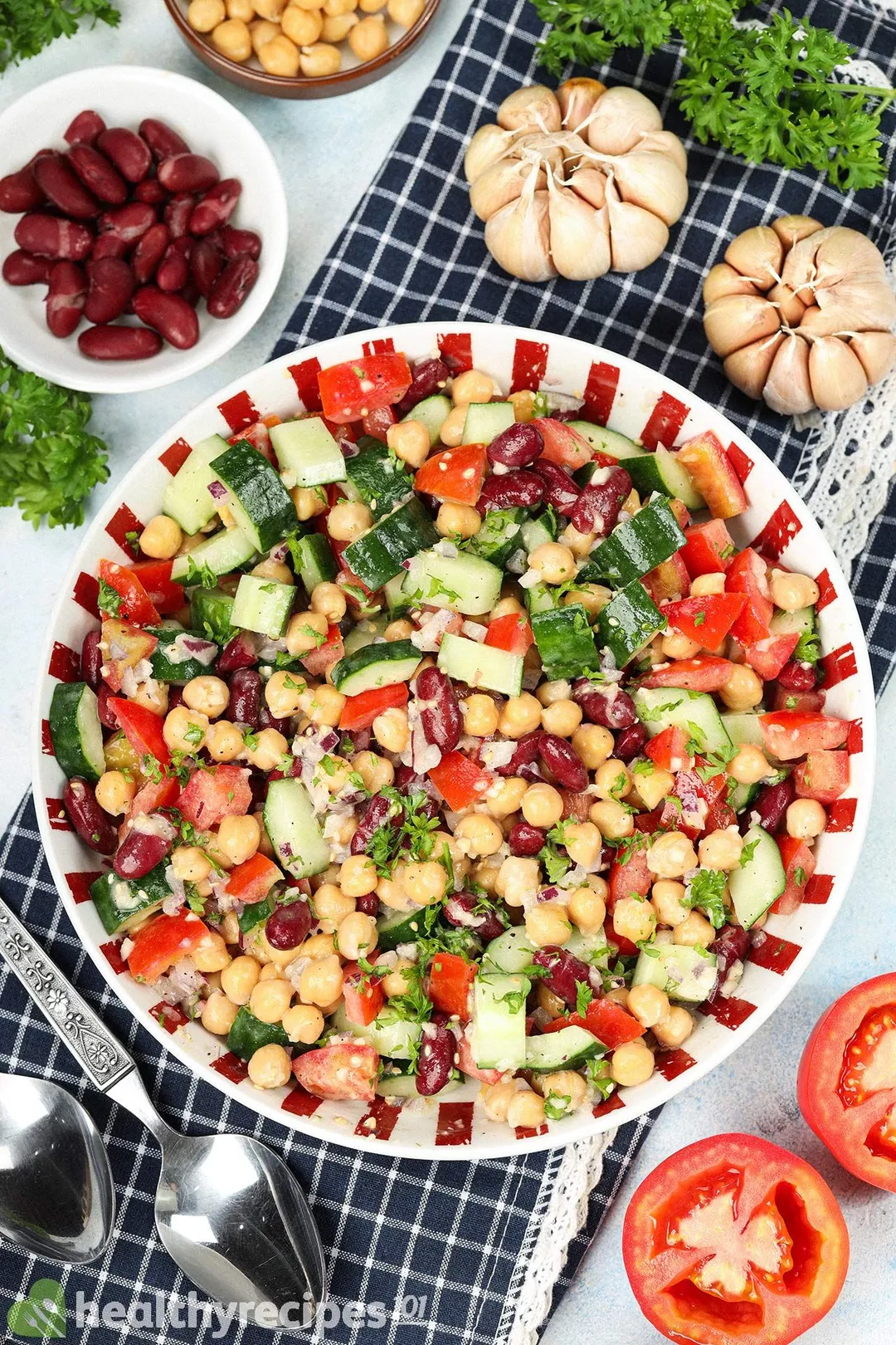 A plate of vegan chickpea salad laid on a blue tablecloth near garlic cloves, kidney beans, spoons, and tomato halves.