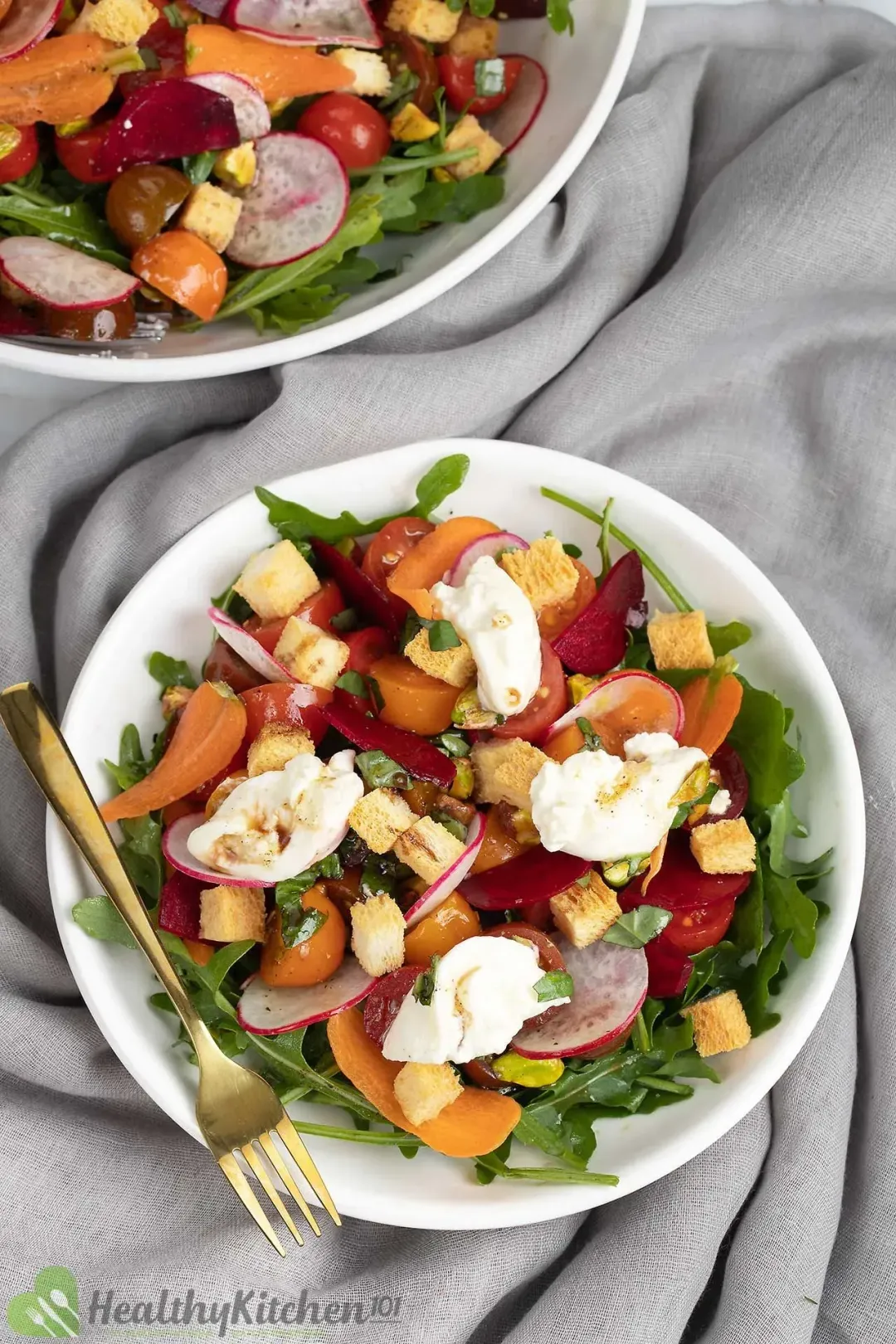 A plate of burrata salad with cherry tomatoes, arugula, carrot shavings, croutons, and burrata slices on a gray cloth