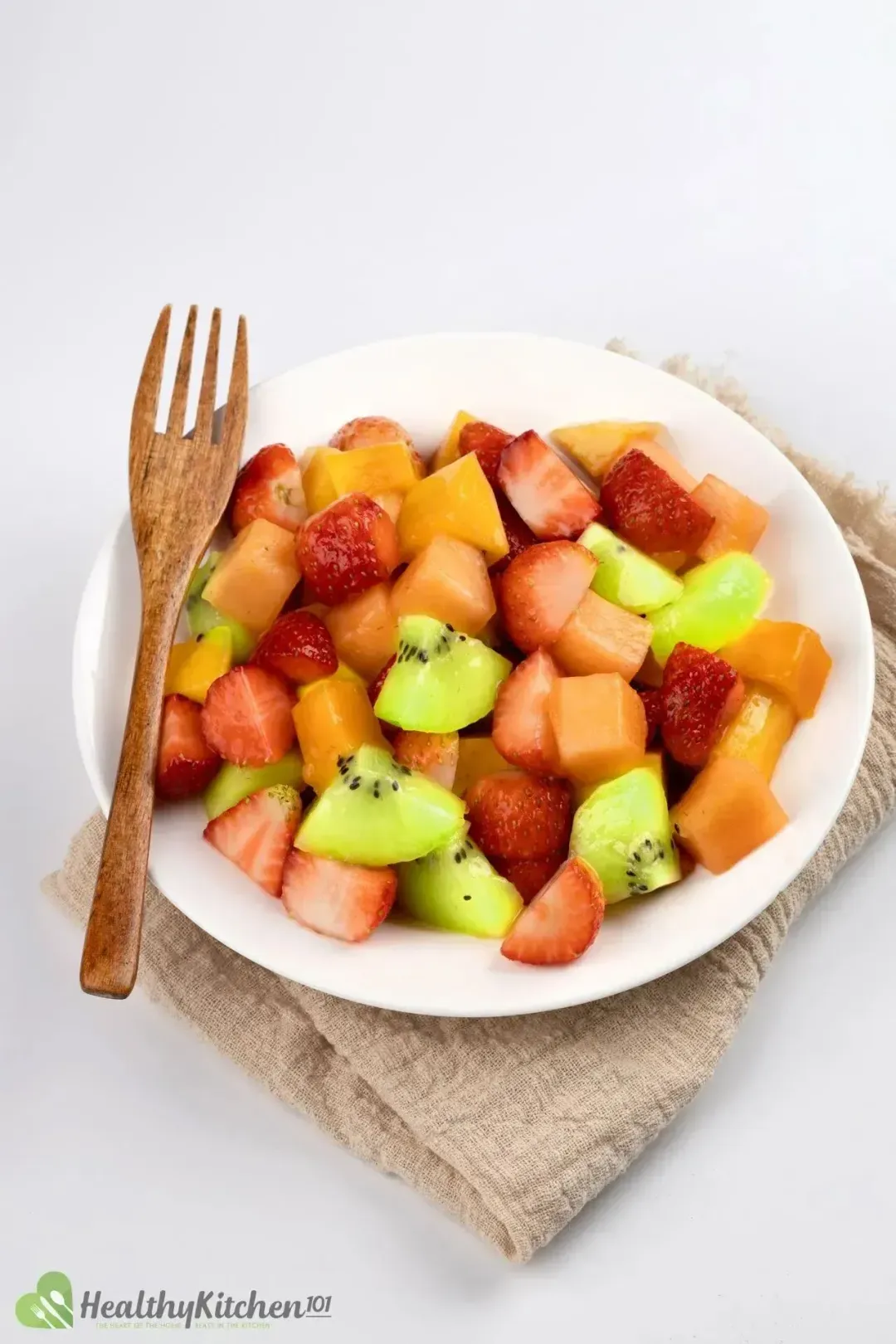 A close-up of a fruit salad in a white bowl with a wooden fork next to it