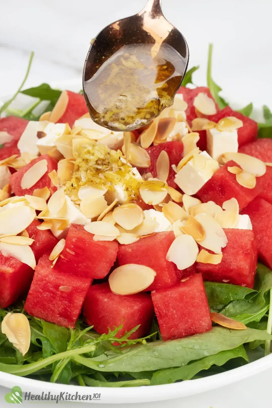 A closeup shot of a salad featuring cubed watermelon, cubed feta, shaved almonds, and arugula, with dressing drizzled on top