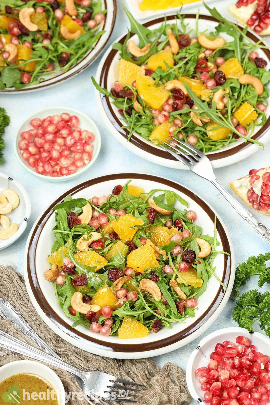 Two plates of Arugula Salad placed near forks and plates of pomegranate seeds.
