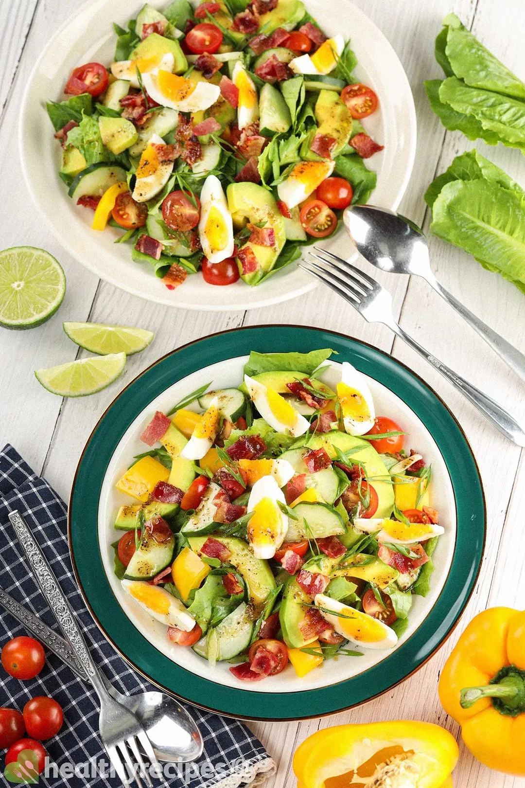 A high-angle shot of two chopped salad plates laid near a blue tablecloth and surrounded by yellow bell peppers, cherry tomatoes, lime wedges, a fork, and a spoon