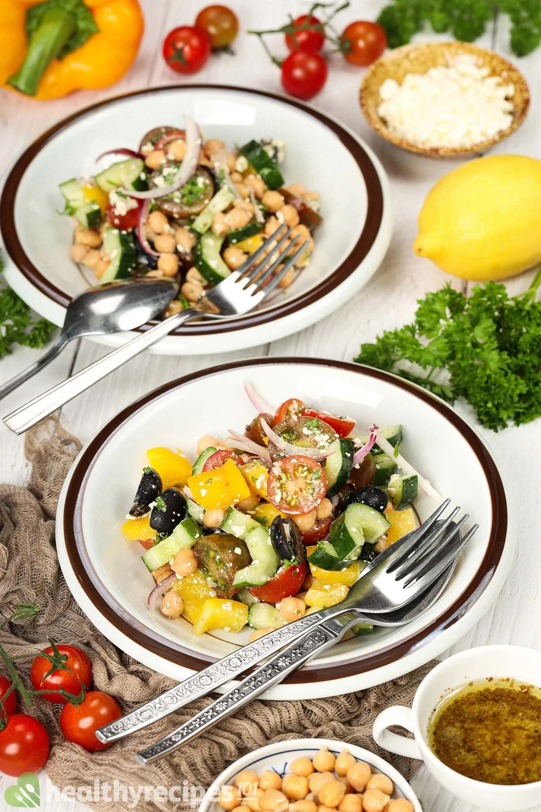 Plates of Greek chickpea salad surrounded by cherry tomatoes, a brown mesh cloth, a lemon, fresh parsley, and a small dressing cup.