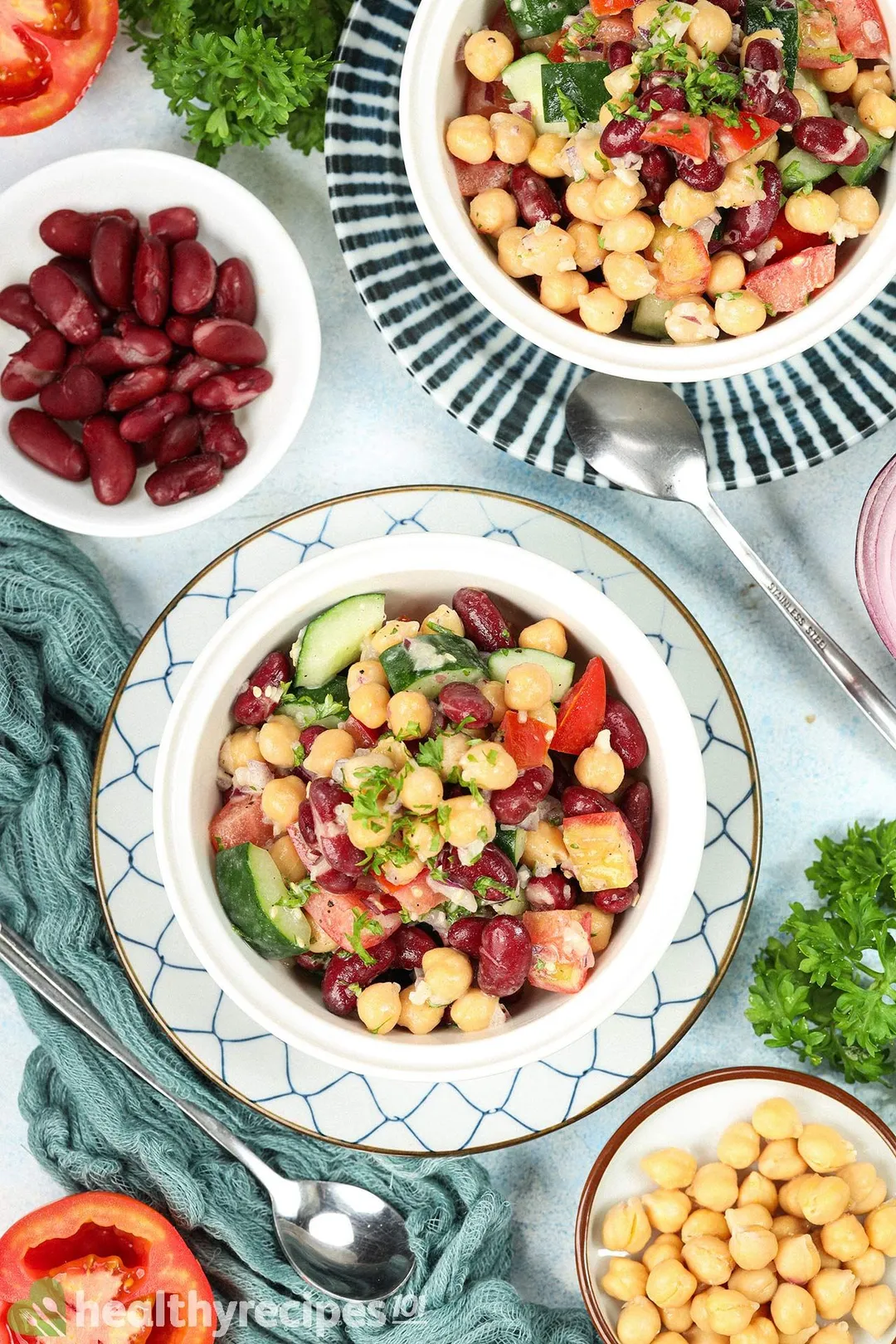 A high-angle shot of two plates of vegan chickpea salad laid next to a small disk of kidney beans, chickpeas, a blue mesh cloth, fresh parsley, spoons, and tomato halves.