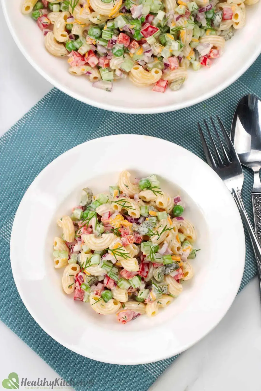 Two white plates of macaroni salad with bits of peas, cheese, dill, pepper, and red onion on a blue mat with silver cutleries