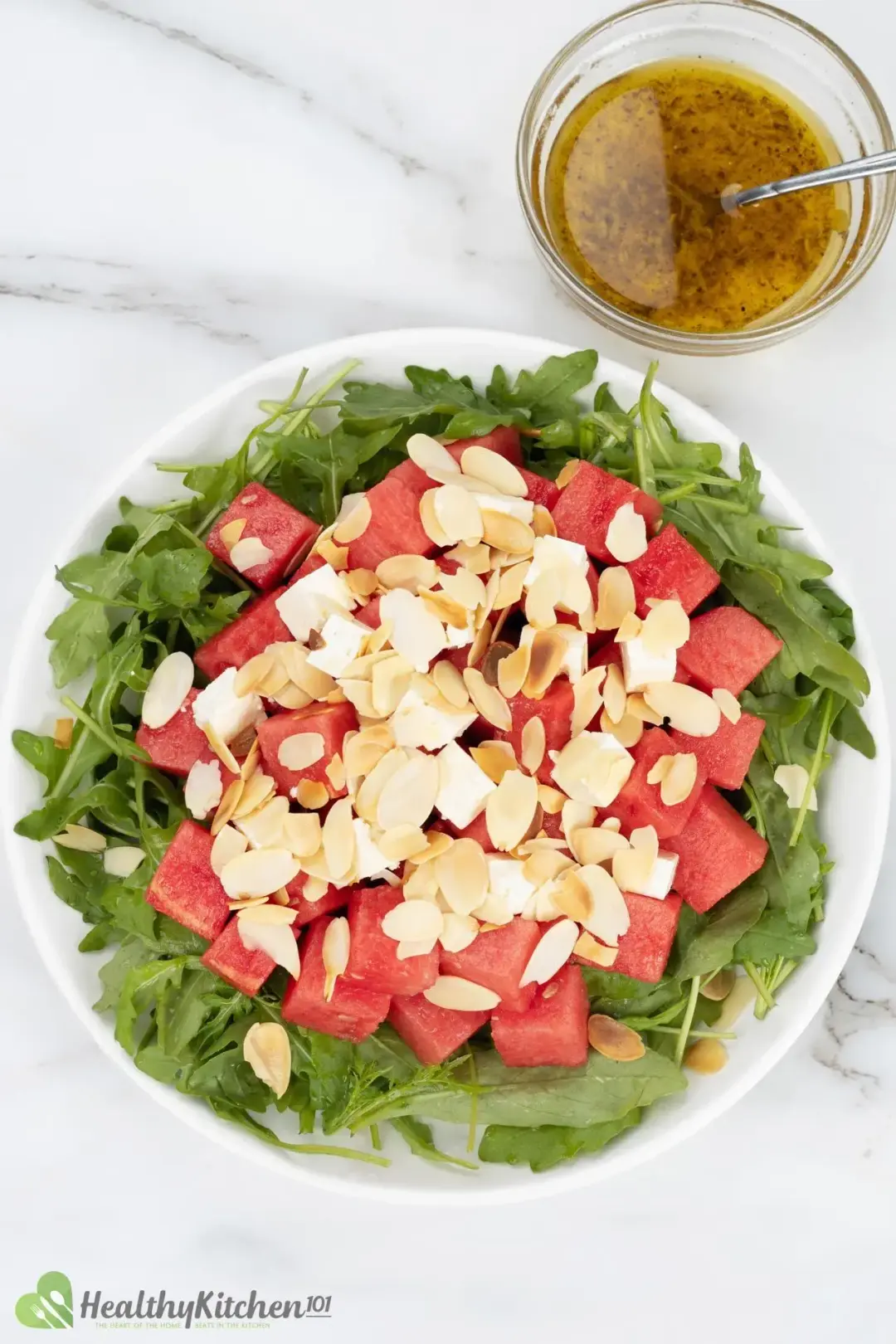 A plate of watermelon cubes and arugula salad, topped with feta and slices of almond, put next to dressing