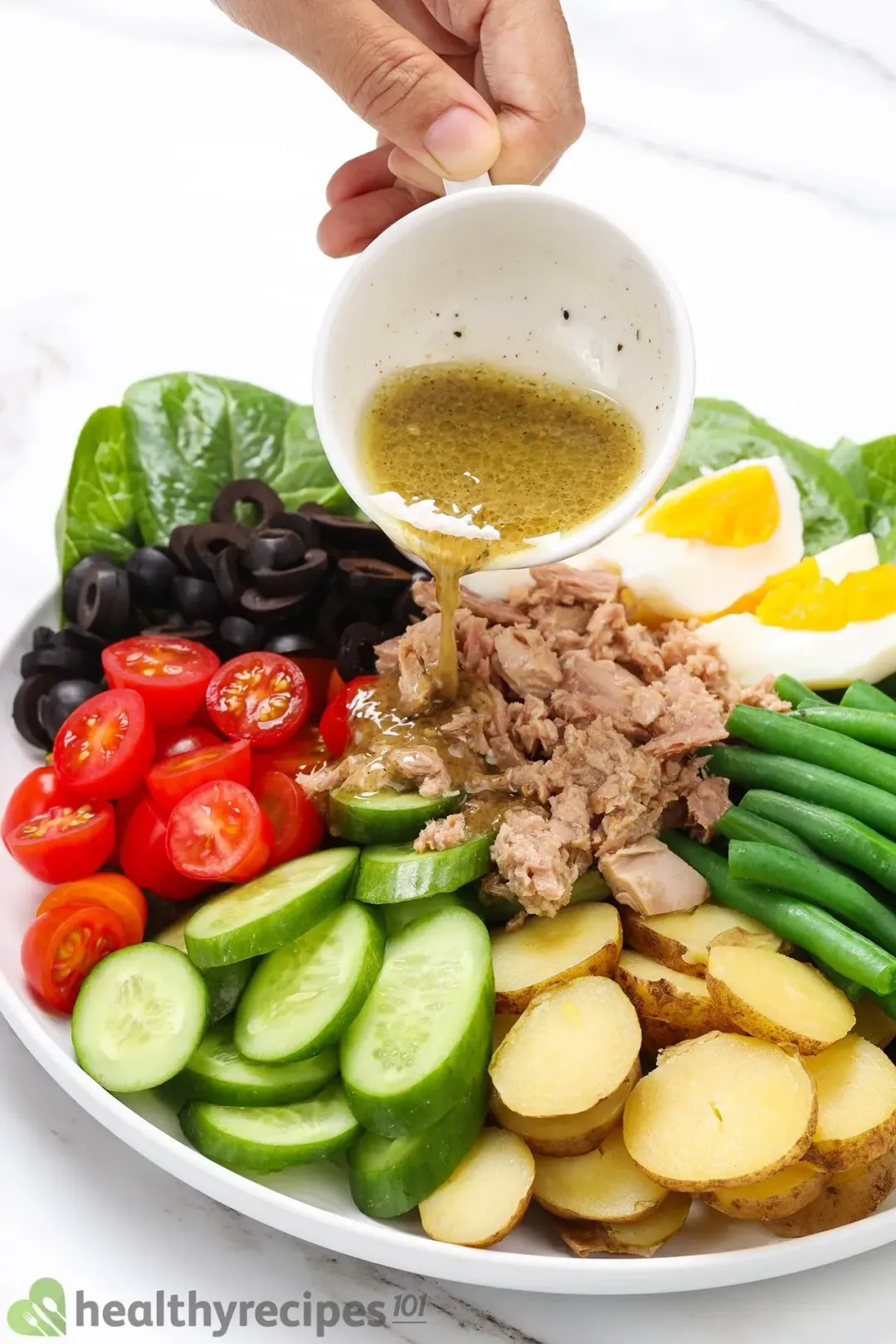 A Nicoise salad with canned tuna, boiled eggs, olives, green beans, tomatoes, and potatoes on a white plate, poured with dressing on top