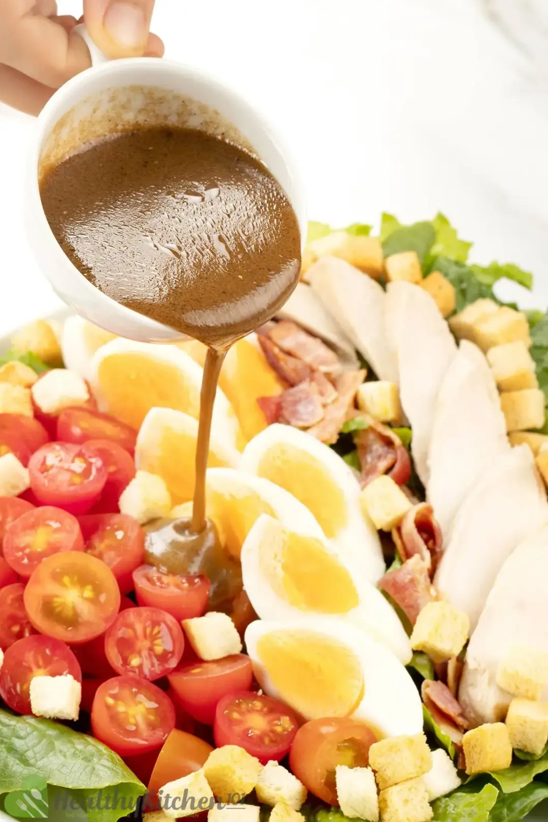 Caesar salad dressing in a glass jar pouring onto a plate of eggs, cherry tomatoes, sliced chicken, and other salad ingredients