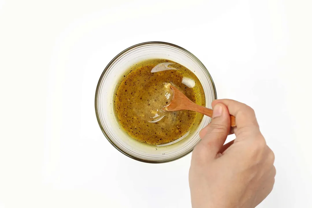 A hand using a wooden spoon to stir a salad dressing in a small bowl.