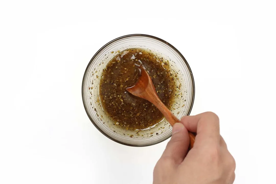 A hand holding a wooden fork stirring around in a small glass bowl containing a dark brown salad dressing