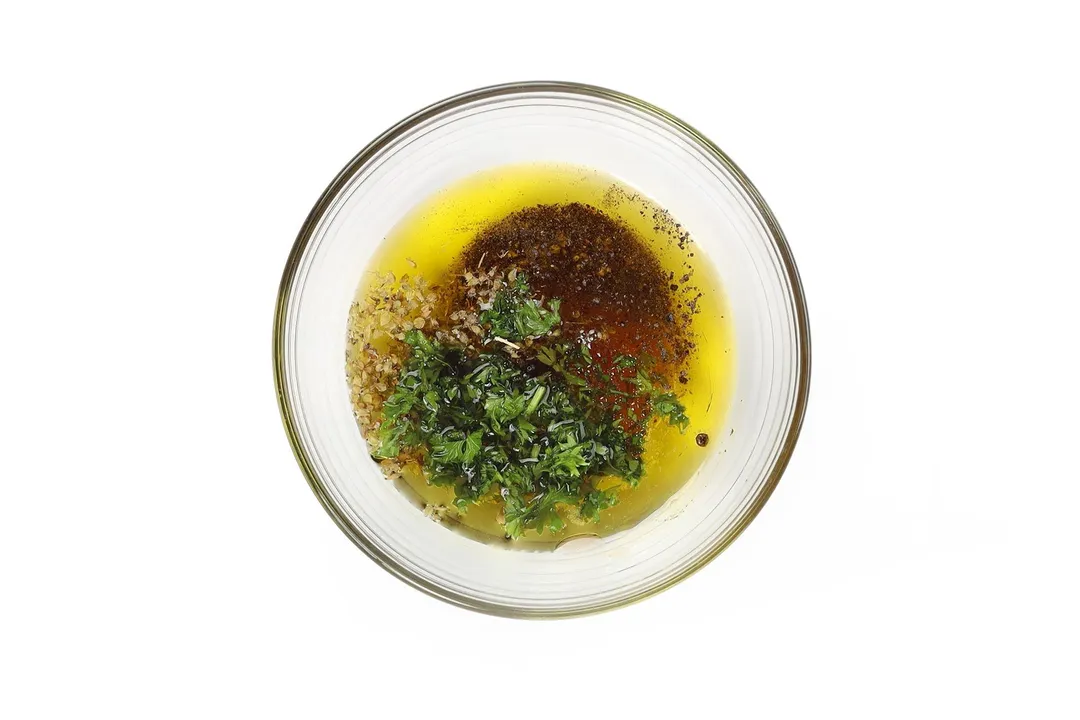 A small glass bowl containing olive oil, spices, and chopped parsley