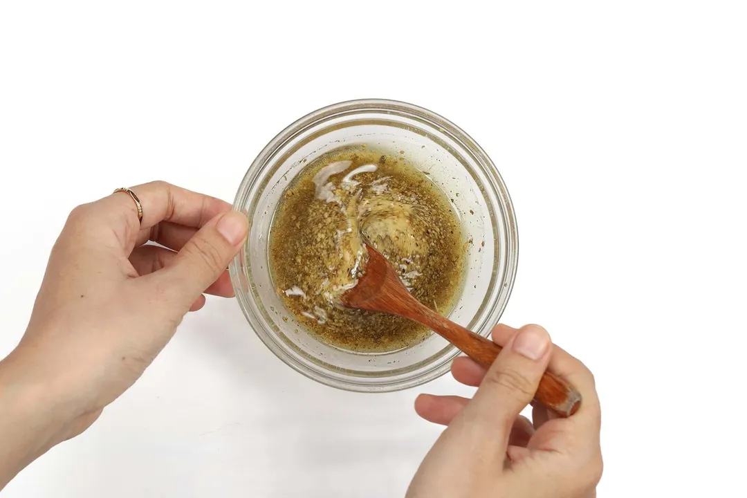 A hand using a wooden spoon to whisk a dark brown salad dressing in a small glass bowl.