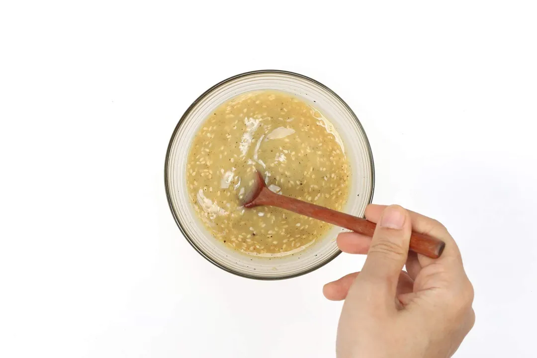 A hand using a wooden spoon to mix a dark yellow mustard dressing in a small glass bowl.