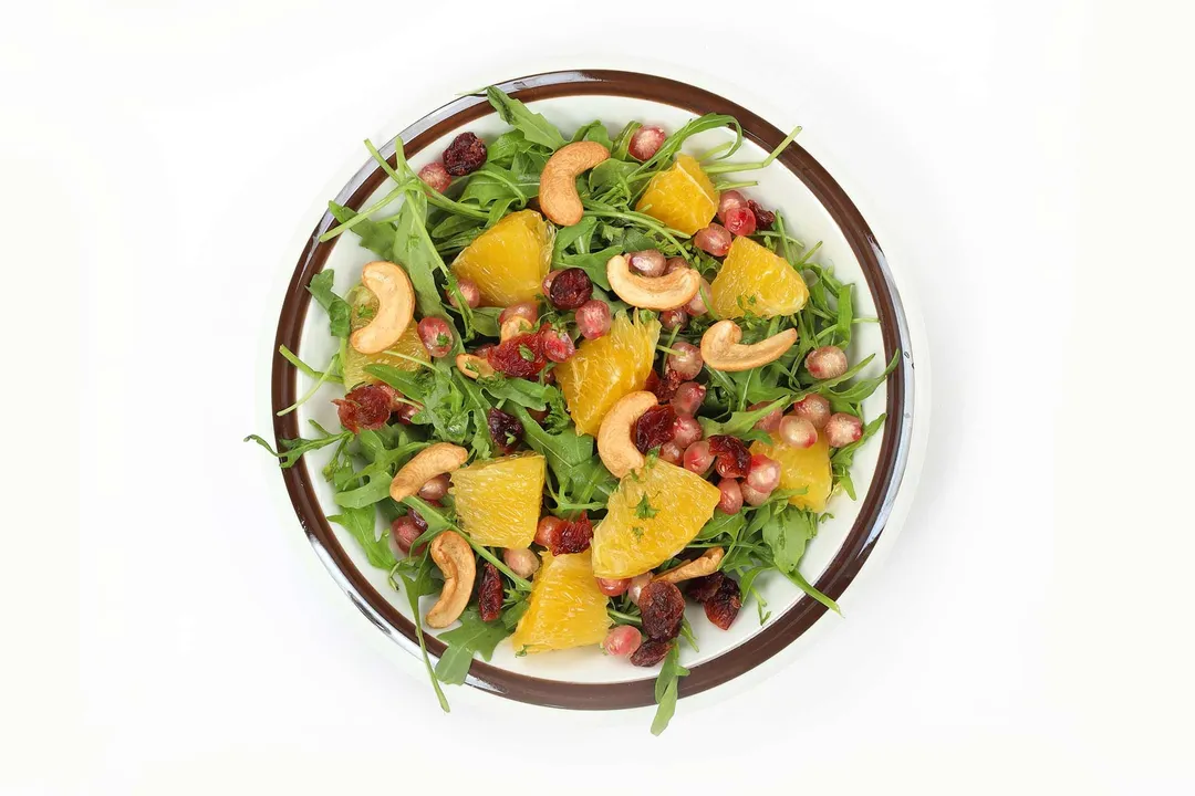 A plate of Arugula Salad including orange segments, arugula leaves, pomegranate seeds, and cashews.