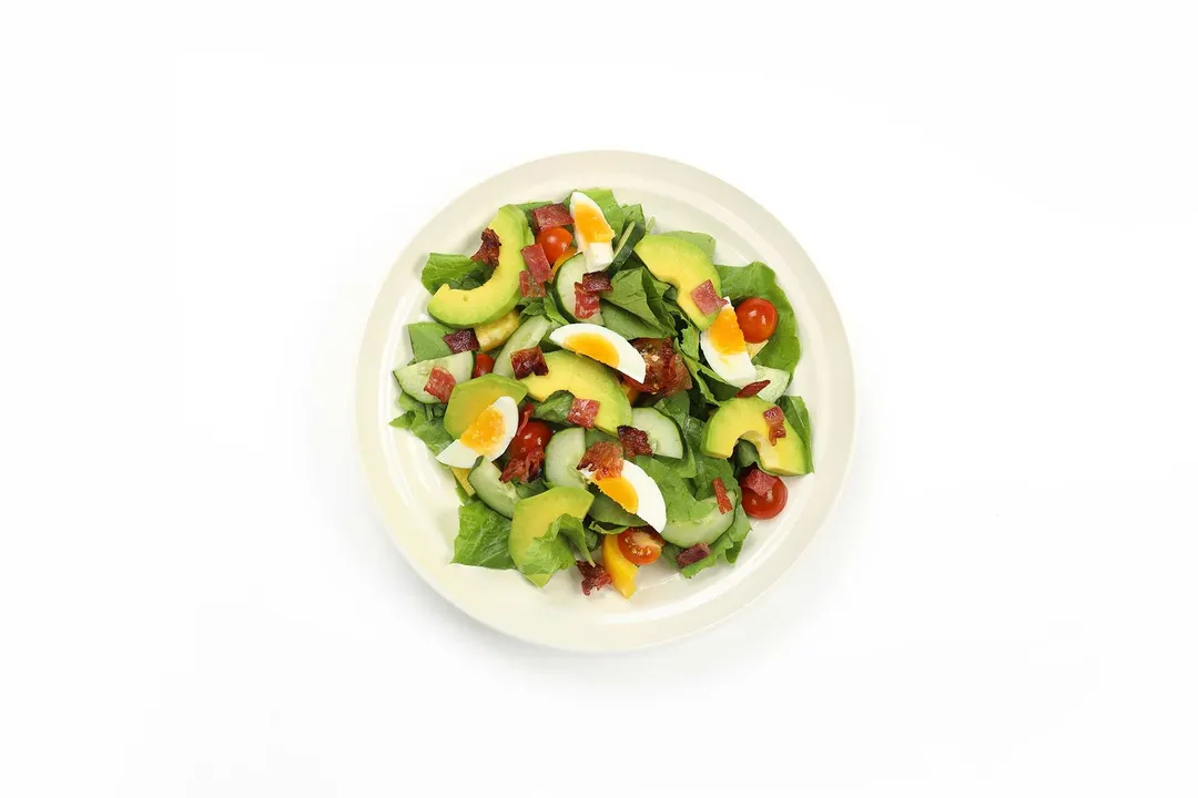 A white plate containing a chopped salad filled with avocado slices, hard-boiled egg wedges, halved cherry tomatoes, and lettuce leaves