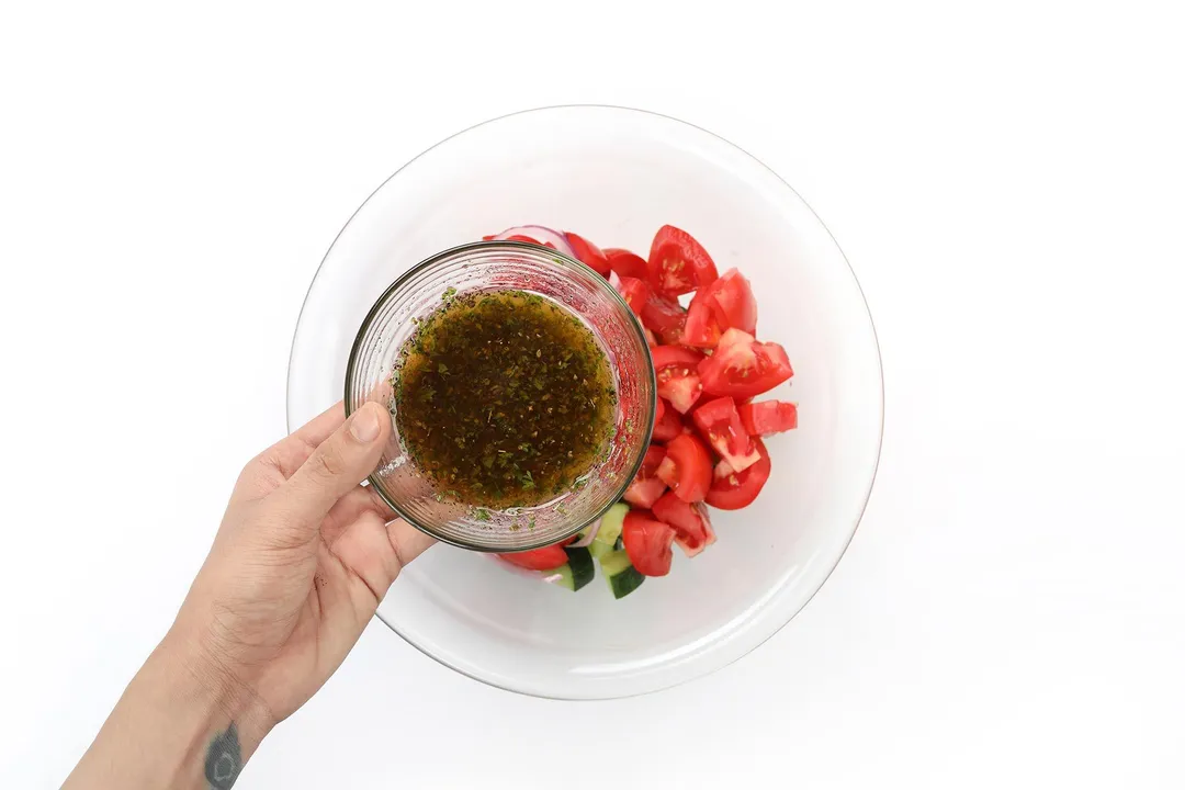 A hand holding a small bowl filled with a dark brown dressing over a large glass bowl containing tomato quarters