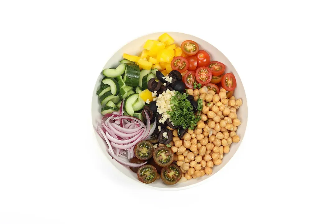 A round white plate with sections of chickpeas, cherry tomato halves, diced bell peppers, sliced cucumbers, sliced red onion, black olives, minced garlic, and chopped parsley on it.
