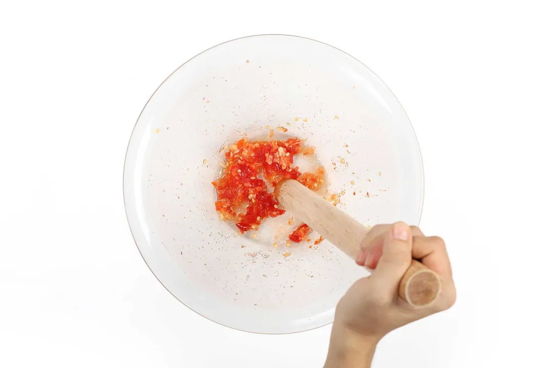 A hand using a wooden pestle to crush a chili into small pieces in a large glass bowl