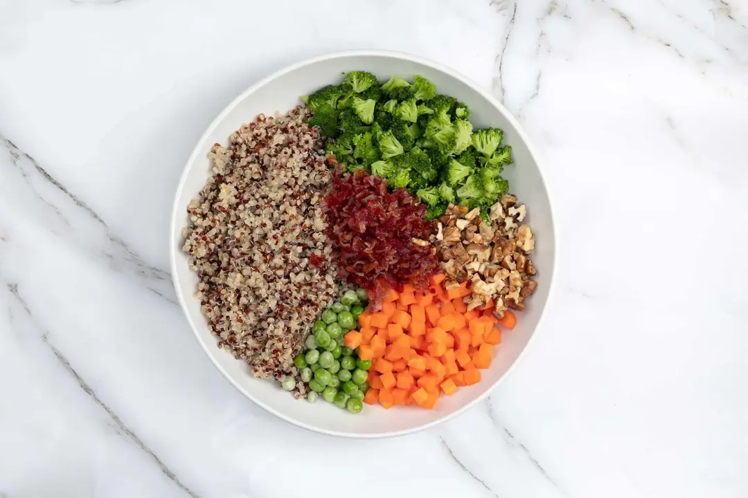 A plate of colorful salad ingredients: quinoa, peas, carrots, broccolis, walnuts, and bacon crumbles