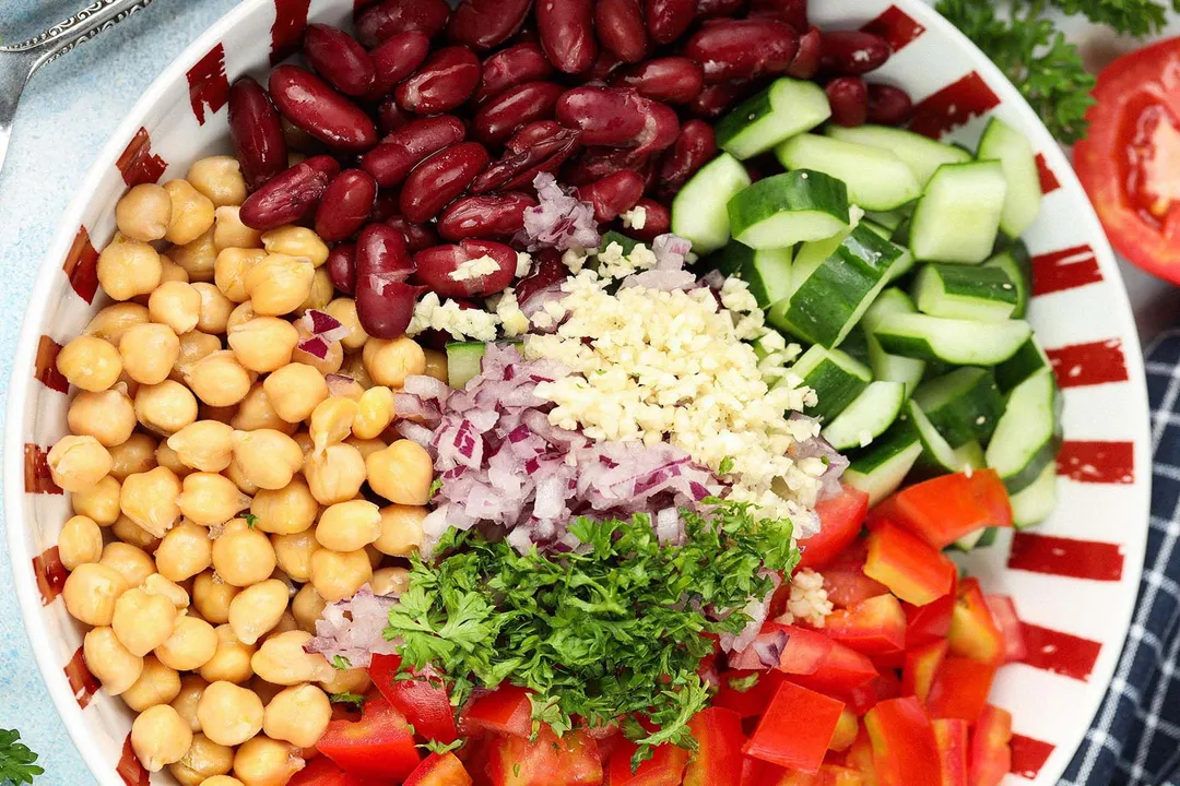 A plate with sections of kidney beans, chickpeas, coarsley-diced cucumbers, coarsley-diced tomatoes, chopped parsley, diced red onion, and minced garlic on it.