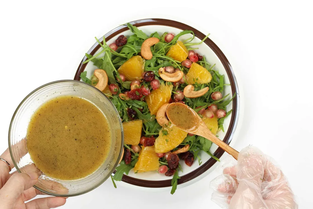 A hand using a wooden spoon to distribute dressing onto a plate of Arugula Salad.