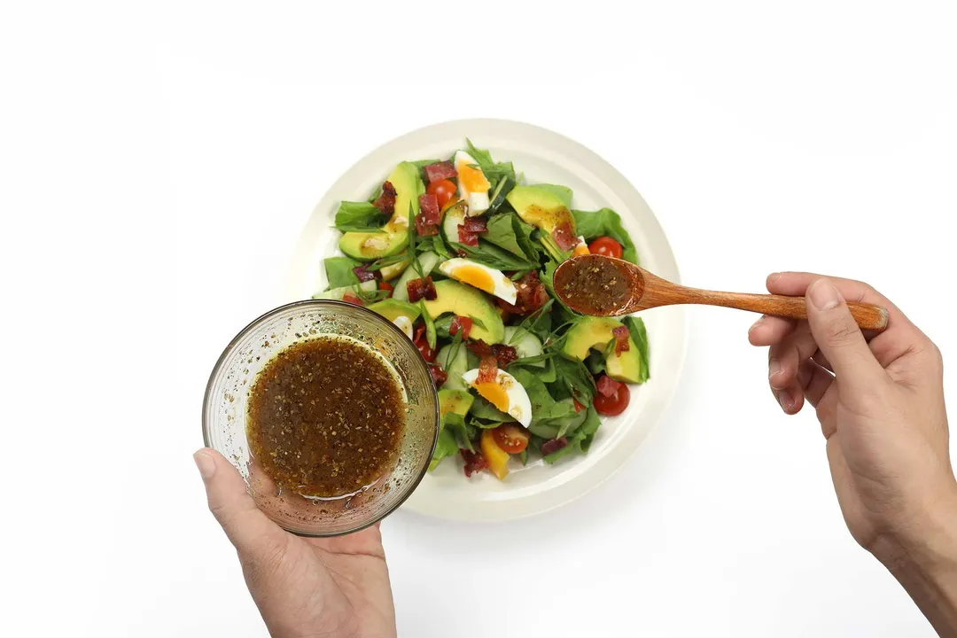 A hand holding a wooden spoon scooping a brown salad dressing onto a plate of chopped salad