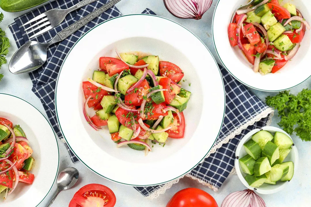 A high-angle shot of cucumber tomato salad plates laid on a blue tablecloth, near a fork, a spoon, and some veggies