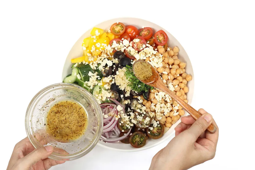 A hand holding a small dressing bowl while the other hand is using a wooden spoon to distribute the dressing onto the plate of Greek chickpea salad beneath it.