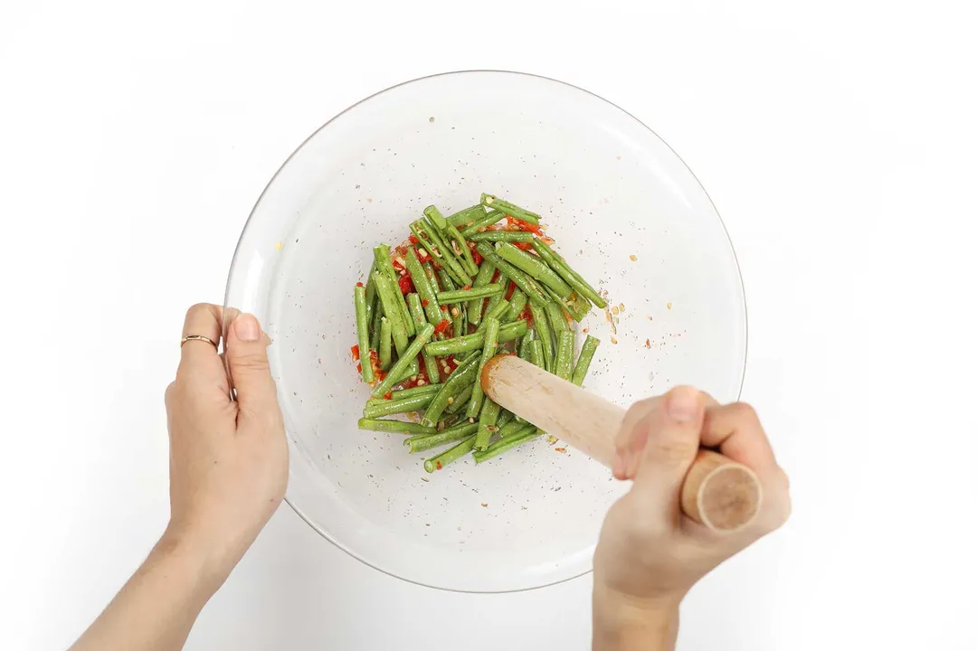 A hand using a wooden pestle to crush a chili and long beans together in a large glass bowl