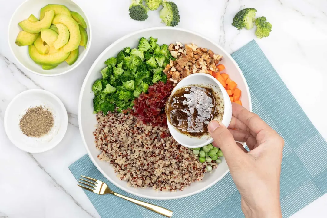 Pouring vinaigrette over a plate of colorful salad ingredients, on a blue mat next to some avocados, peppers, and broccoli florets