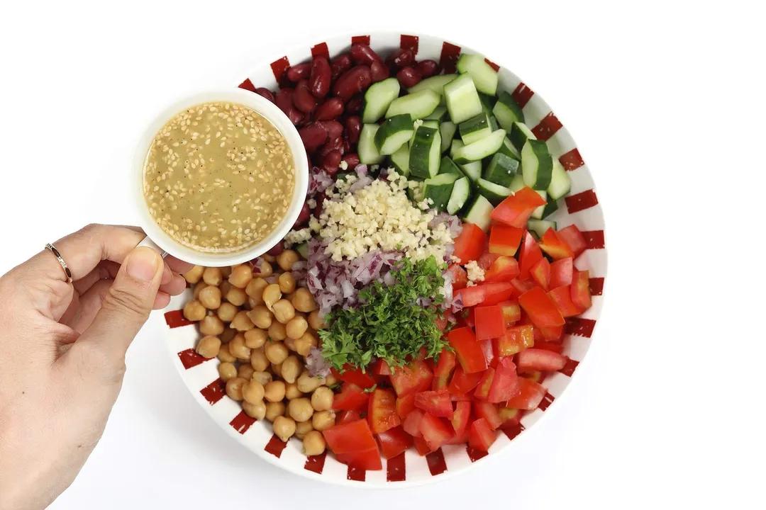 A hand holding a small bowl of dressing to drizzle over a plate filled with sections of kidney beans, tomatoes, cucumbers, parsley, onion, and garlic.