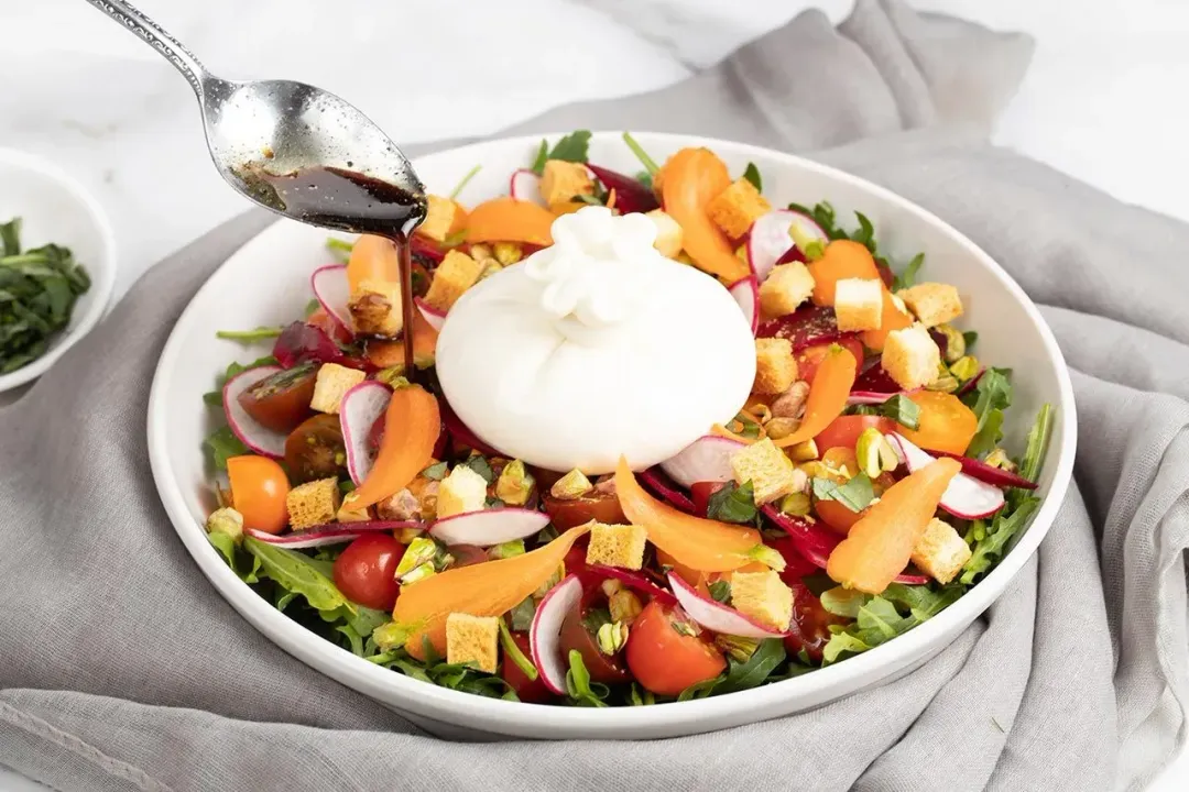 Burrata salad with arugula, cherry tomatoes, croutons, carrot shavings, and dressing being added from a spoon