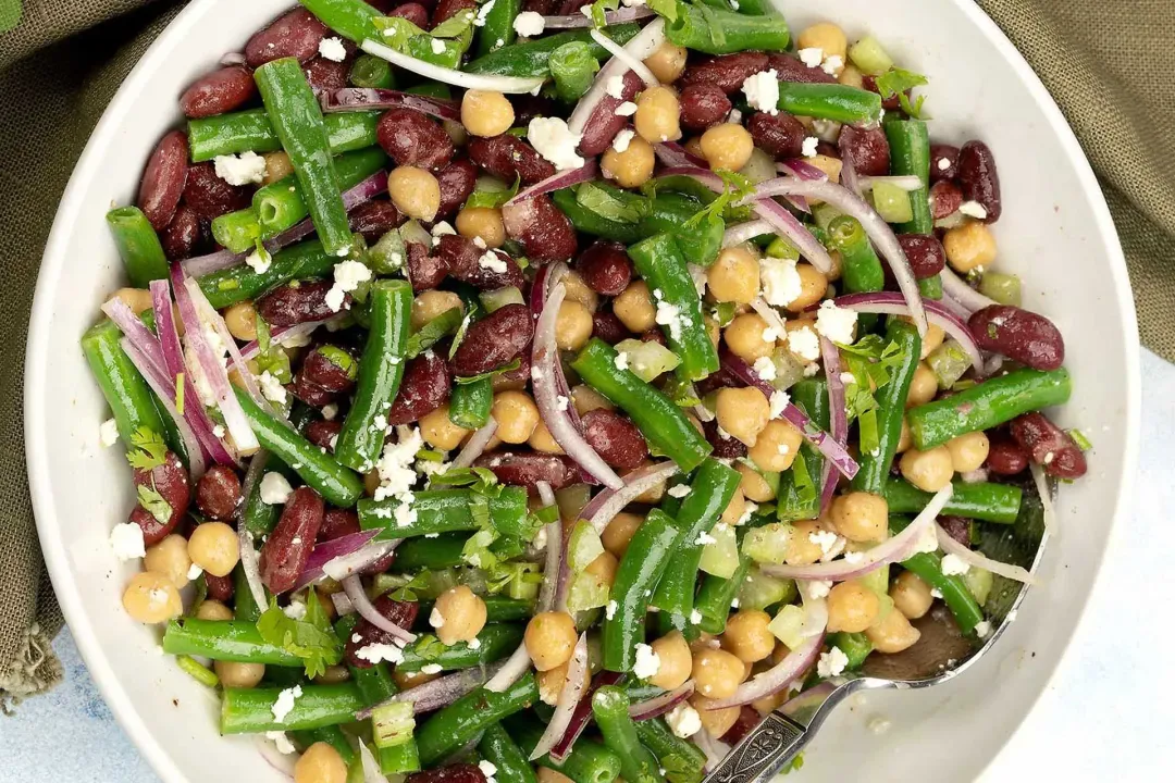 A bowl of three-bean salad with dressing, red onion slices, and cheese cubes