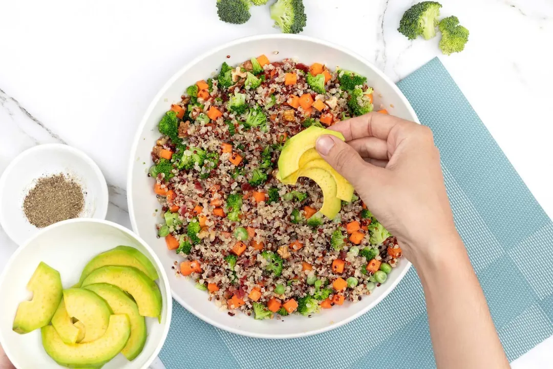 Placing avocado slices on a plate of quinoa salad with broccoli, carrot, next to some broccoli florets and avocado slices