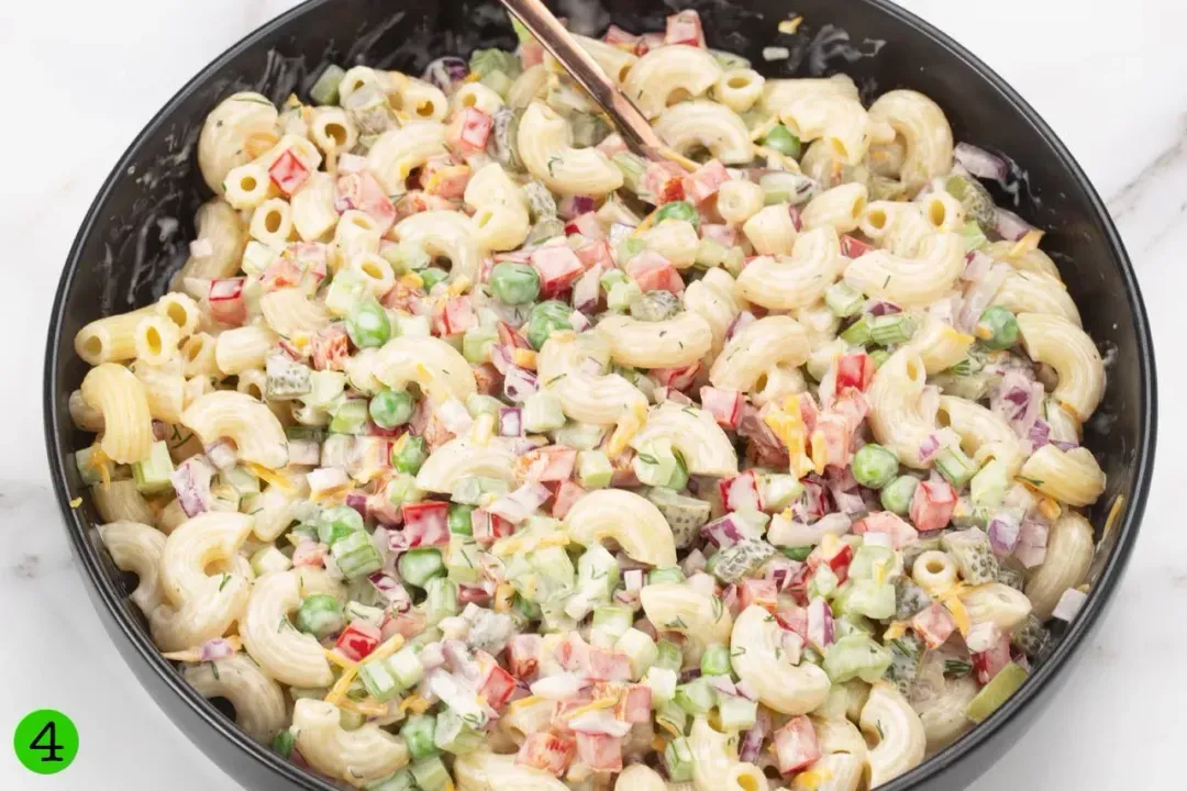 A bowl of macaroni salad with cooked macaroni, pepper, cheese, peas, pepper, and mayo dressing tossed together.