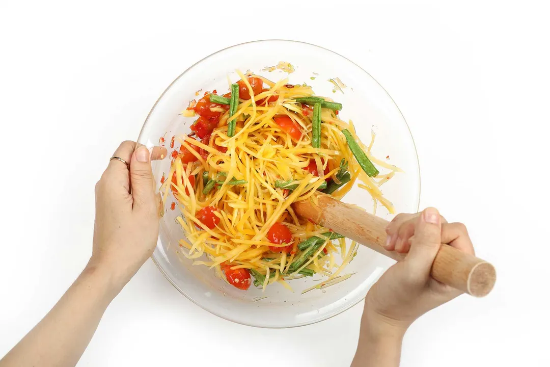 A hand using a wooden pestle to mix shredded papaya, halved tomatoes, and long beans together in a large glass bowl