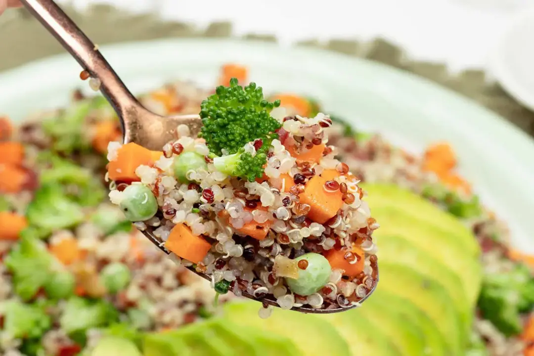 A spoon full of quinoa carrot pea broccoli salad pieces, hovering on a plate of quinoa salad and avocado slices
