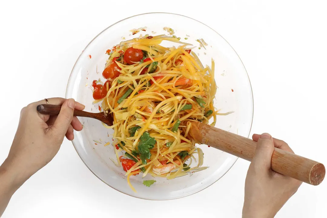 Hands using a wooden pestle and a wooden spoon to mix shredded papaya, halved tomatoes, and long beans together in a large glass bowl
