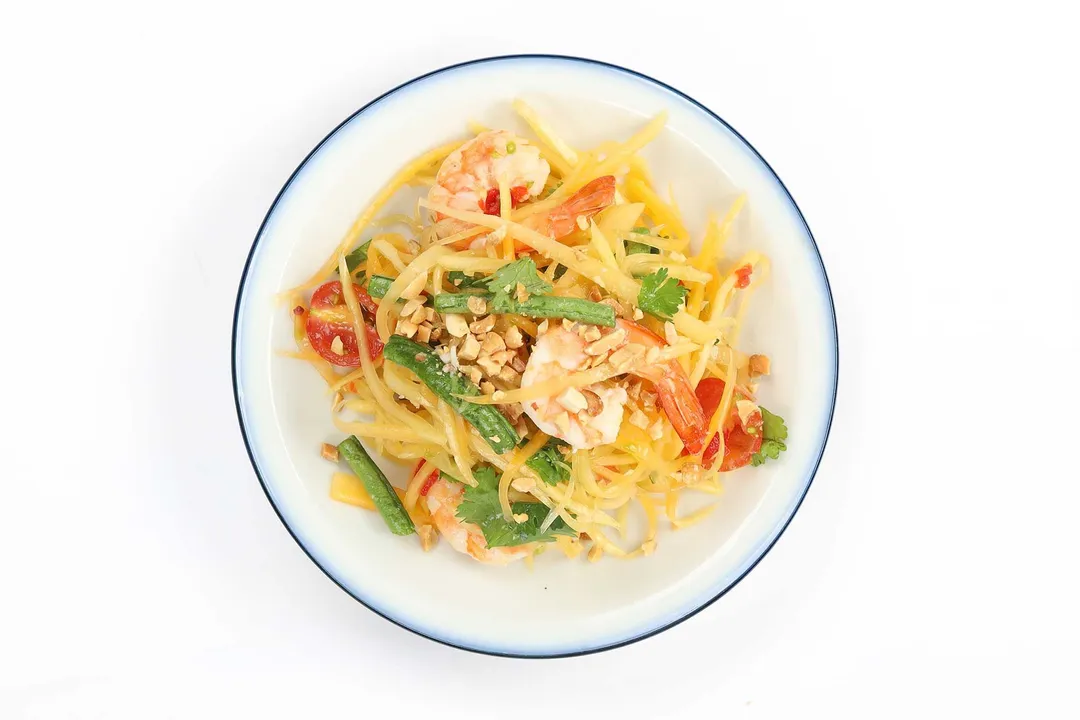 Papaya salad consisting of shredded papaya, cooked shrimp, long beans, and roasted peanuts laid on a round white plate with blue rim
