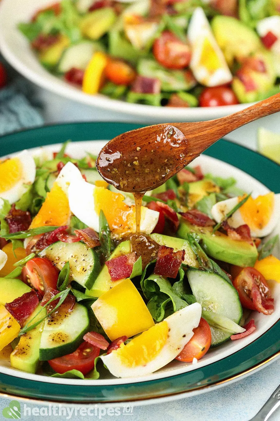 A wooden spoon distributing a thick dressing onto a chopped salad plate filled with hard-boiled eggs, halved cherry tomatoes, sliced cucumbers, and lettuce