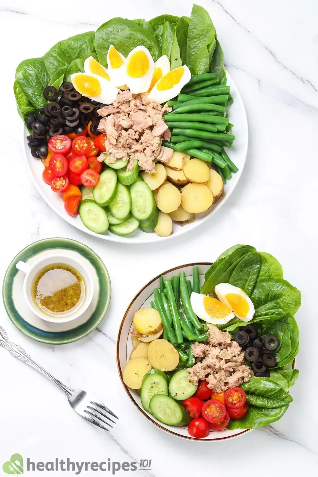 Two plates of Nicoise salad with tuna, eggs, olives, tomatoes, potatoes, green beans, and cucumbers, next to a fork and dressing