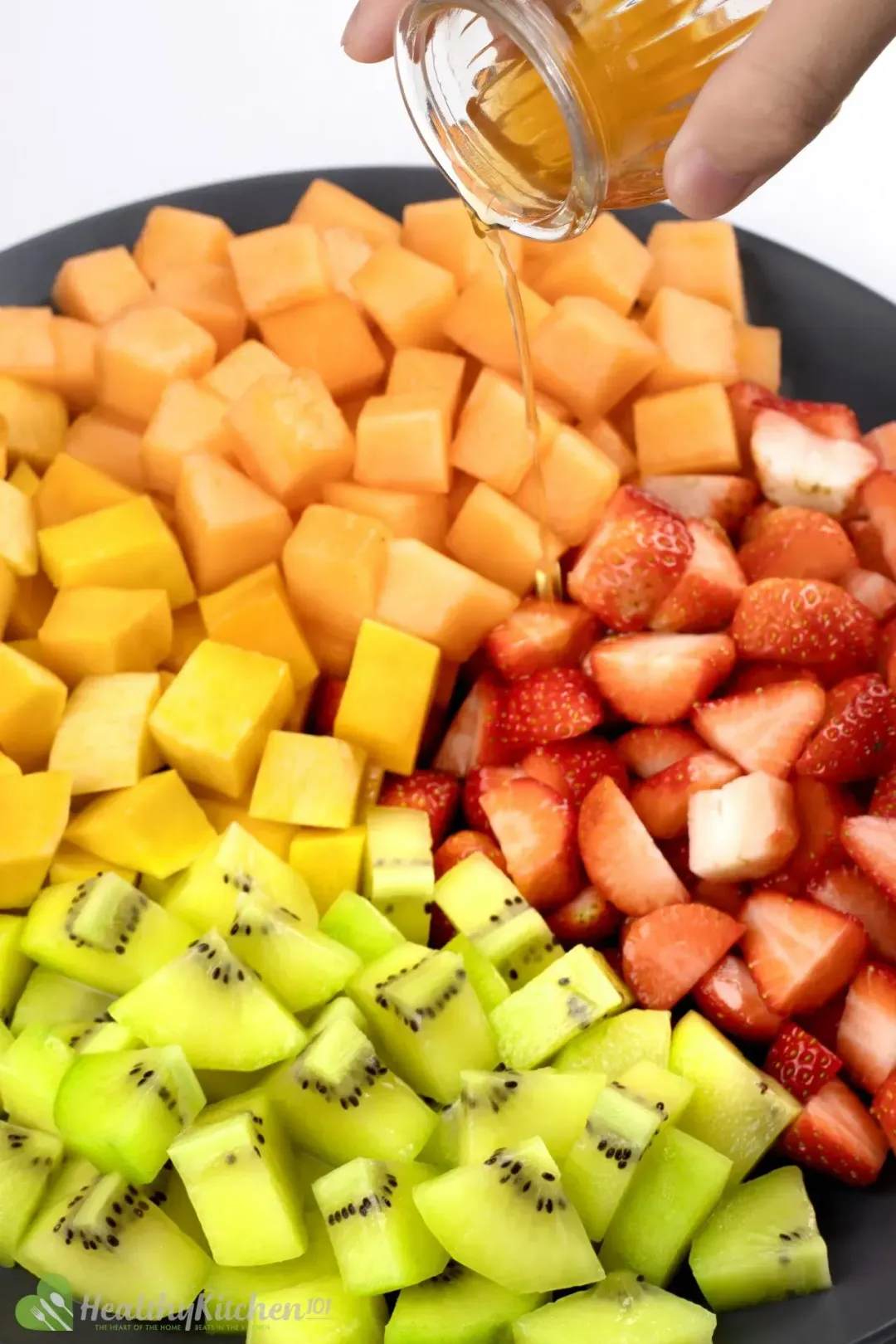 A colorful fruit salad with strawberries, blueberries, kiwi, and oranges on a white plate, being poured on top with honey