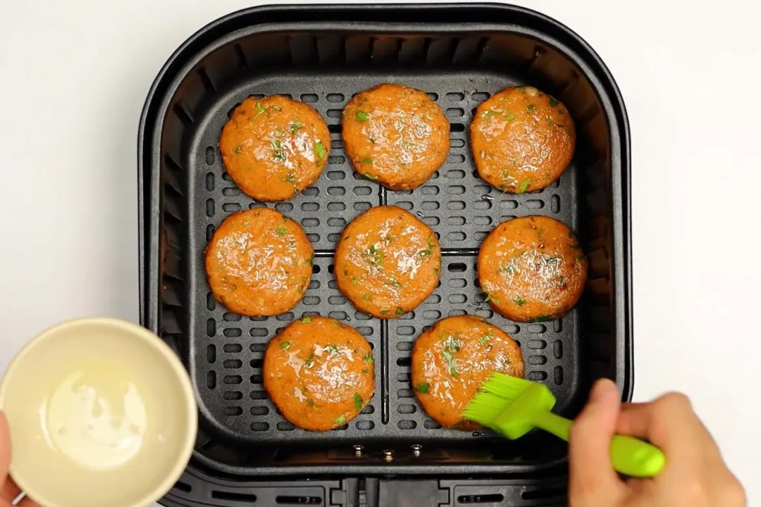 A hand is brushing some olive oil over eight salmon patties placed in an air fryer’s basket.